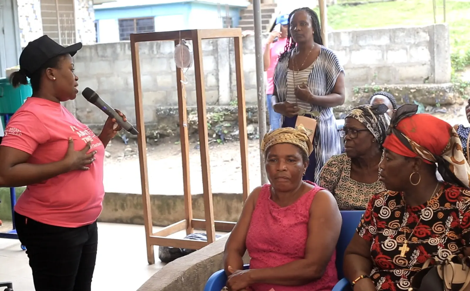 A Stepin team member in a pink T-shirt speaks into a microphone to women seated at an outdoor community cancer education session.