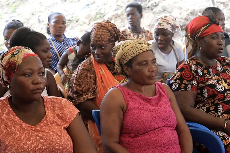 Women in colourful headwraps and dresses sit together and listen during a community outreach session.