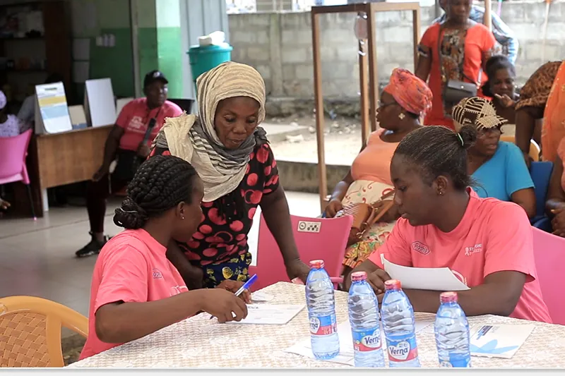 Two Stepin team members in pink T-shirts write on forms at a table while a woman in a headscarf stands beside them at a community outreach.