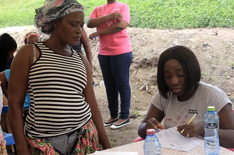 A woman in a striped top stands beside a seated woman who is filling in a form at an outdoor table, with a person in a pink T-shirt standing behind.